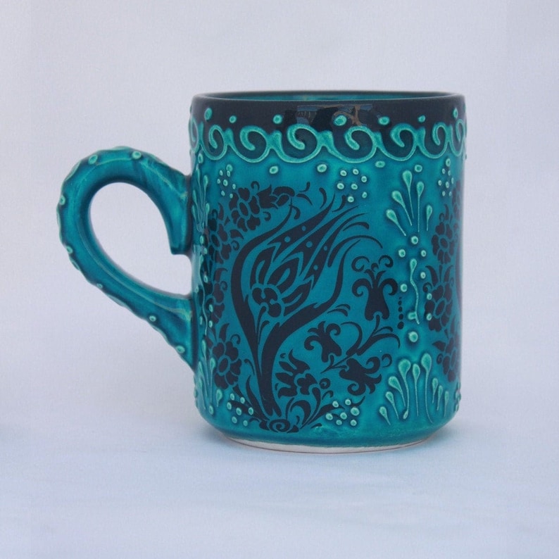 Puede incluir: Una taza de cer&aacute;mica turquesa con un dise&ntilde;o floral negro. La taza tiene un asa y un borde negro.