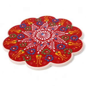 May include: Red ceramic coaster with a white and multi-colored floral design. The coaster has a scalloped edge.