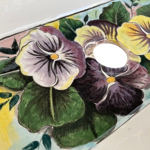 May include: A white ceramic sink with a hand-painted design of purple, yellow, and green pansies. The sink has a drain hole in the center.