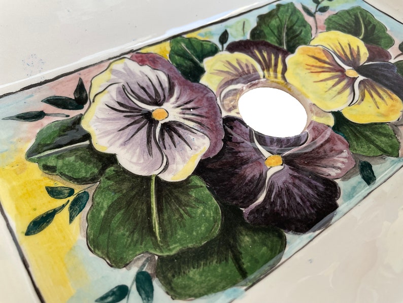 May include: A white ceramic sink with a hand-painted design of purple, yellow, and green pansies. The sink has a drain hole in the center.