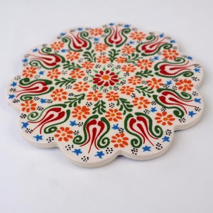 May include: Round ceramic trivet with a white background and a colorful floral design. The design features red, orange, green, and blue tulips, flowers, and leaves.