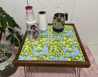 May include: A wooden table with a blue and yellow floral design. The table has a metal frame and is decorated with a vase, a mug, and a small ceramic container.