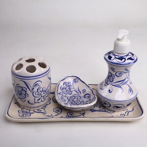 May include: A white ceramic bathroom set with blue floral designs. The set includes a toothbrush holder, a soap dispenser, a soap dish, and a tray.