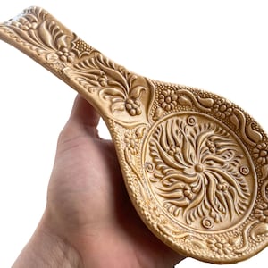May include: A gold ceramic spoon rest with an intricate floral design. The spoon rest is shaped like a spoon and has a raised rim to catch drips.