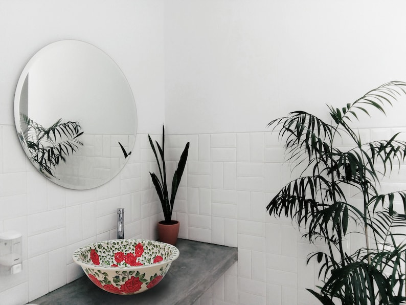 May include: A white ceramic bathroom sink with a floral design featuring red roses and green leaves. The sink is in a modern bathroom with white tile walls and a round mirror.