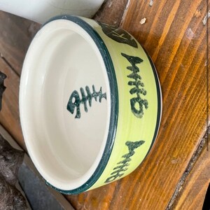 Pu&ograve; includere: Una piccola ciotola per animali domestici in ceramica gialla con un bordo verde e un disegno di lisca di pesce nero. La ciotola dice "Whiskers" sul lato.