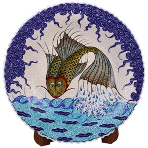 Op de afbeelding: Een wit keramiek bord met een blauw en gouden visontwerp. De vis springt uit het water, dat in blauw en groen is afgebeeld. Het bord heeft een gegolfde rand en is versierd met blauwe swirls.