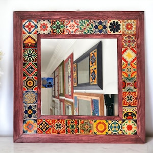 Puede incluir: Un espejo cuadrado con un marco de madera decorado con azulejos de mosaico de colores. El marco es de color burdeos oscuro y los azulejos son una mezcla de rojo, amarillo, azul, verde y negro.