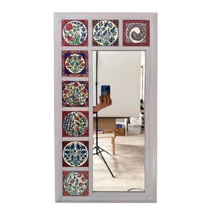 Puede incluir: Un espejo con marco de madera blanco con un espejo rectangular rodeado de ocho azulejos cuadrados con motivos florales coloridos. Los azulejos están dispuestos en dos columnas de cuatro azulejos cada una.