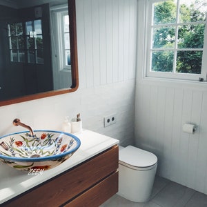May include: A white ceramic bathroom sink with a floral design in shades of blue, red, and green. The sink is in a modern bathroom with white walls and a wooden vanity.