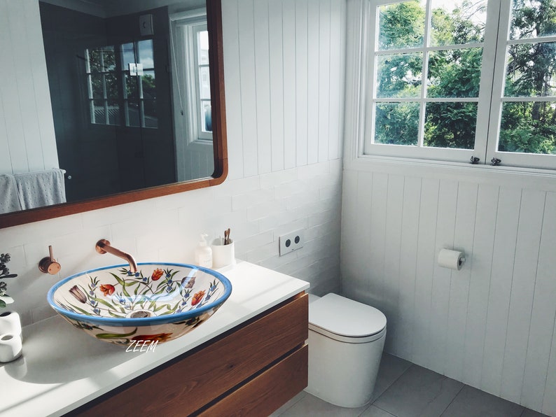 May include: A white ceramic bathroom sink with a floral design in shades of blue, red, and green. The sink is in a modern bathroom with white walls and a wooden vanity.