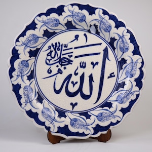 Puede incluir: Un plato de cerámica blanco con un diseño floral azul y blanco alrededor del borde. El centro del plato presenta una inscripción caligráfica azul de la palabra "Allah" en árabe.