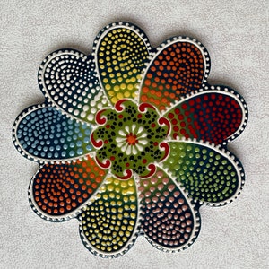 May include: A colourful ceramic trivet shaped like a flower with a central design of dots and swirls. The trivet is decorated with a range of colours, including red, orange, yellow, green, blue, and purple.