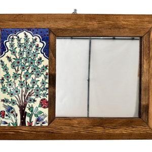 Puede incluir: Espejo rectangular con marco de madera y una inserción de azulejo de cerámica decorativo. El azulejo presenta un diseño de árbol con flores azules y turquesas, detalles rojos y un borde azul. El espejo es rectangular.