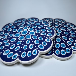 Puede incluir: Dos posavasos de cerámica azul y blanco con un patrón de ojos azules. Los posavasos tienen forma de flor con bordes redondeados.
