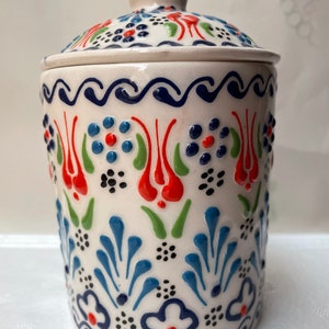 Op de afbeelding: Witte keramische pot met deksel, versierd met een blauw, rood en groen bloemenpatroon. De pot heeft een traditioneel Turks ontwerp.