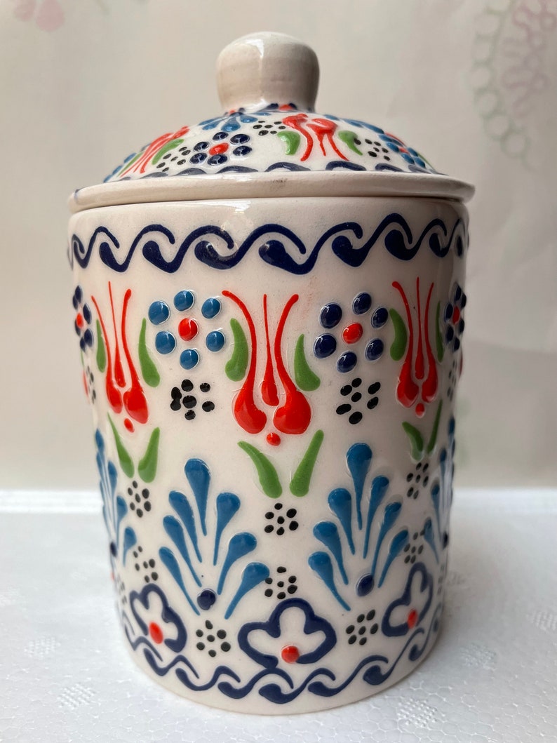 Op de afbeelding: Witte keramische pot met deksel, versierd met een blauw, rood en groen bloemenpatroon. De pot heeft een traditioneel Turks ontwerp.