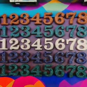 May include: Five rows of ceramic number tiles, each row featuring the numbers 0 through 9. The tiles are decorated with a floral pattern and are arranged in a rainbow of colors: brown, teal, white, blue, and green.
