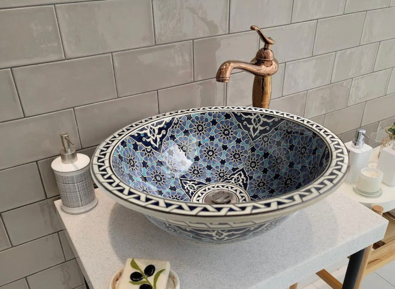 Pu&ograve; includere: Un lavabo da bagno in ceramica blu e bianca con un motivo floreale decorativo. Il lavabo ha un bordo bianco e uno scarico al centro. Il lavabo &egrave; su un piano di lavoro bianco con uno schienale in piastrelle grigie.