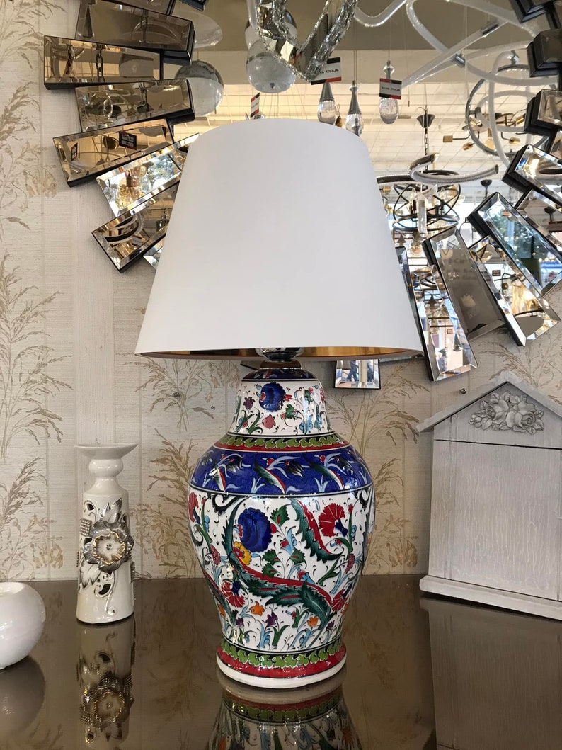 Peut inclure: Une lampe de table en c&eacute;ramique blanche avec un abat-jour blanc. Le pied de la lampe pr&eacute;sente un motif floral color&eacute; dans les tons bleu, vert, rouge et jaune.
