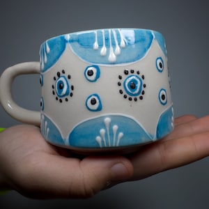Op de afbeelding: Een witte keramische mok met een blauw-wit ontwerp. De mok heeft een handvat en is versierd met een patroon van blauwe ogen en witte stippen.