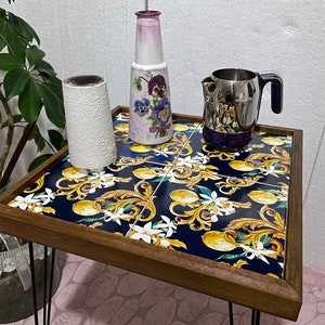 Puede incluir: Una mesa auxiliar de madera con patas de metal negro. La superficie de la mesa es una baldosa de cerámica con fondo azul y un patrón de limones, flores y volutas doradas.