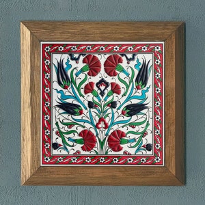 Może przedstawiać: Płytka ceramiczna w ramie z tradycyjnym tureckim wzorem. Płytka ma białe tło z czerwonymi, niebieskimi i zielonymi wzorami kwiatowymi.