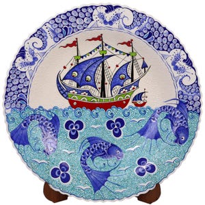 Op de afbeelding: Een rond keramiek bord met een blauwe en witte rand. Het midden van het bord heeft een blauw en rood schip met witte zeilen, omringd door blauwe vissen op een turquoise achtergrond.