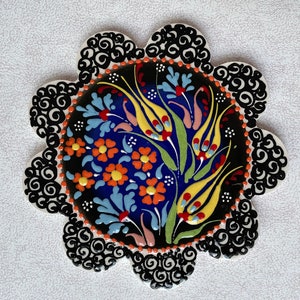 Könnte beinhalten: Eine dekorative Keramikfliese mit schwarzem Hintergrund und einem farbenfrohen Blumendesign. Das Design zeigt gelbe, blaue, rote, orangefarbene und grüne Blumen und Blätter. Die Fliese hat einen gewellten Rand mit einem schwarz-weißen Muster.