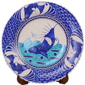 Op de afbeelding: Een blauw en wit keramisch bord met een visontwerp. Het bord heeft een gegolfde rand en is versierd met een vis die in het water zwemt. De vis wordt omringd door blauwe en witte golven en schubben. Het bord staat op een houten standaard.