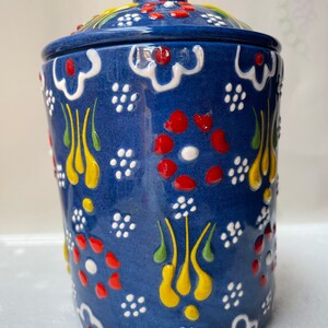 Op de afbeelding: Een blauwe keramische pot met een deksel, versierd met witte, rode, gele en groene bloemmotieven. De pot heeft een traditioneel Turks ontwerp.