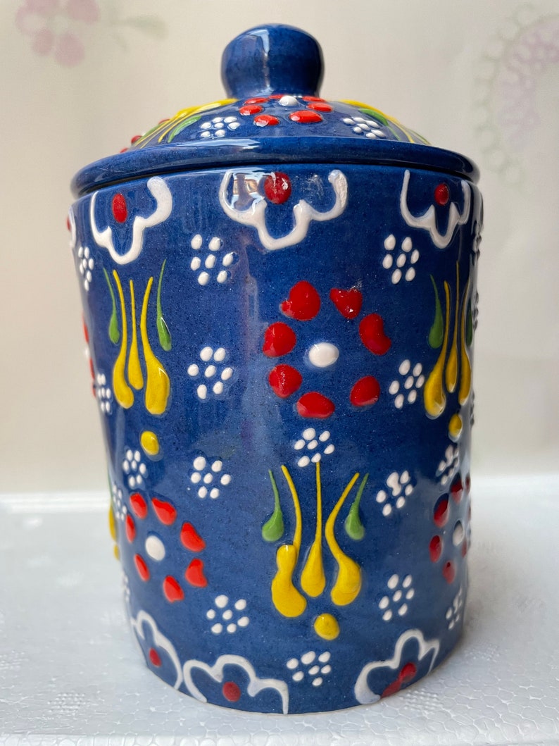 Op de afbeelding: Een blauwe keramische pot met een deksel, versierd met witte, rode, gele en groene bloemmotieven. De pot heeft een traditioneel Turks ontwerp.