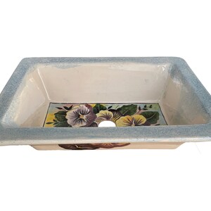 May include: A white ceramic sink with a blue rim and a painted floral design featuring purple and yellow pansies.