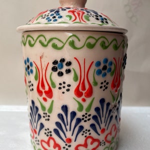 Op de afbeelding: Een witte keramische pot met een deksel, versierd met een kleurrijk bloemenpatroon in tinten rood, blauw, groen en zwart. De pot heeft een traditioneel Turks ontwerp.