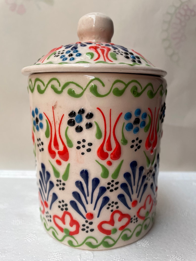 Op de afbeelding: Een witte keramische pot met een deksel, versierd met een kleurrijk bloemenpatroon in tinten rood, blauw, groen en zwart. De pot heeft een traditioneel Turks ontwerp.