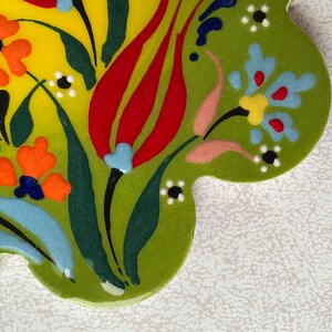 May include: A green ceramic tile with a floral design. The tile features red, yellow, orange, blue, and white flowers on a green background.