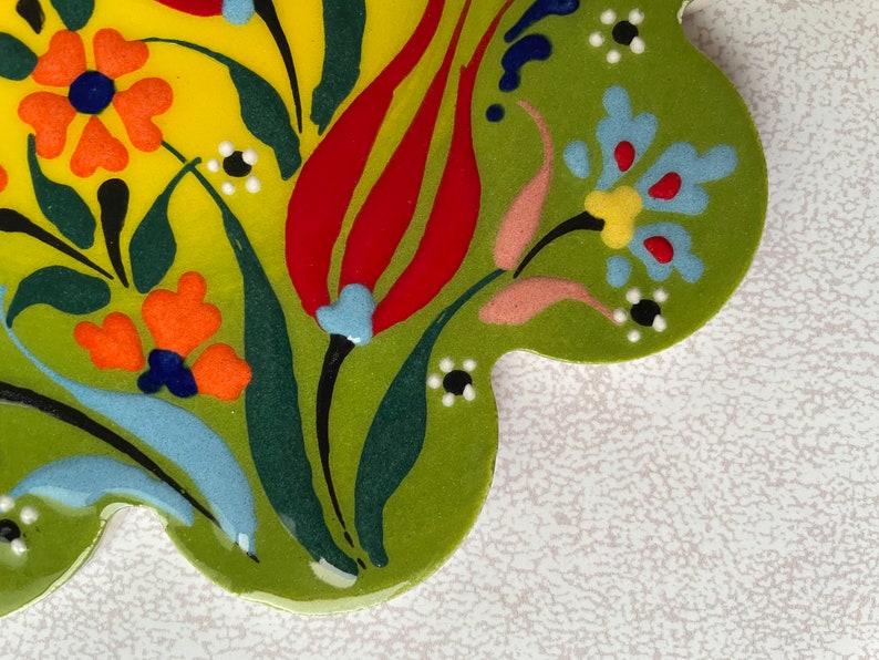 May include: A green ceramic tile with a floral design. The tile features red, yellow, orange, blue, and white flowers on a green background.