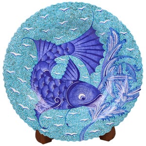 Op de afbeelding: Een blauw en wit keramiek bord met een gedetailleerd ontwerp van een vis die zwemt in een zee van golven. Het bord staat op een houten standaard.