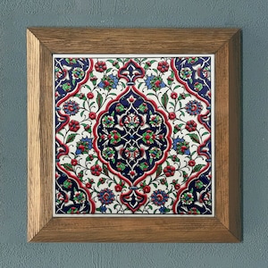 May include: Square ceramic tile with a floral pattern in red, blue, and green, set in a wooden frame. The intricate design features stylized flowers and leaves on a white background, creating a decorative wall art piece.