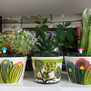 May include: Three ceramic flower pots with different plant designs. The pots are white with green, red, blue, and yellow painted details. The plants are a cactus, a succulent, and a leafy plant.