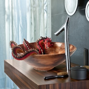 May include: A decorative bathroom sink with a wooden bowl and a chrome tap. A red and brown dragon sculpture is inside the bowl. The sink is on a dark wood vanity with a brush and other accessories.