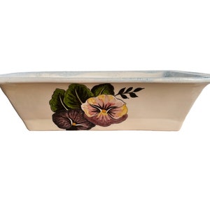 May include: A white ceramic baking dish with a floral design featuring purple and pink pansies with green leaves. The dish has a slightly raised rim.