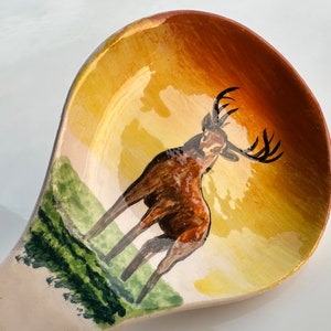 May include: A ceramic spoon with a hand-painted design of a brown deer with antlers standing in a field against a yellow and orange sunset background.