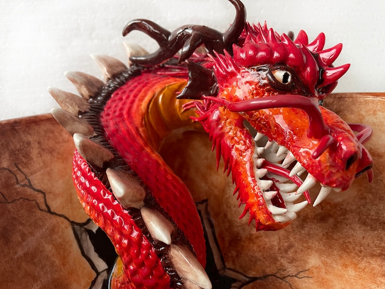 May include: A detailed sculpture of a red and orange dragon's head with sharp white teeth and a long red tongue. The dragon has dark brown horns and a textured, scaly appearance. The sculpture appears to be emerging from a cracked surface.