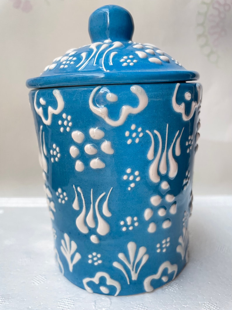 Op de afbeelding: Een blauwe keramische pot met een deksel, met een wit bloemenpatroon. De pot heeft een ronde vorm en een gladde, glanzende afwerking.