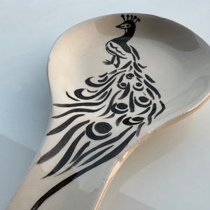 May include: A white ceramic spoon rest featuring a black and white peacock design. The peacock is facing right and has a crown on its head.