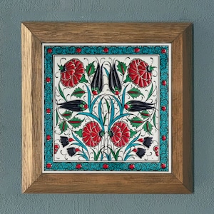 Puede incluir: Un azulejo de cerámica enmarcado con un diseño tradicional turco que presenta flores rojas, tulipanes negros y hojas verdes sobre un fondo blanco. El azulejo está rodeado por un borde turquesa con un patrón repetitivo.
