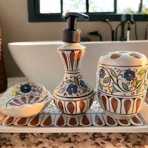 Puede incluir: Una bandeja de cerámica blanca con un diseño floral marrón y blanco contiene un juego de accesorios de baño. El juego incluye un dispensador de jabón, un portacepillos de dientes y un plato pequeño. Todas las piezas están decoradas con motivos florales azules y amarillos.