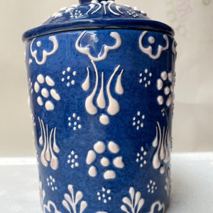 Op de afbeelding: Een blauwe keramische pot met een wit deksel en witte bloemmotieven. De pot heeft een traditioneel Turks ontwerp.