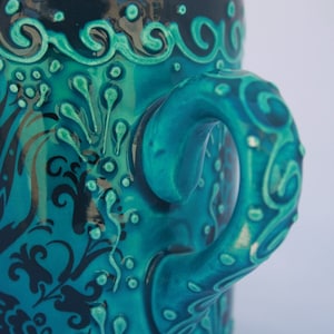 Puede incluir: Una taza de cer&aacute;mica azul turquesa con un dise&ntilde;o floral negro y un asa &uacute;nica. El asa tiene forma de cuerda trenzada y presenta un dise&ntilde;o en relieve.